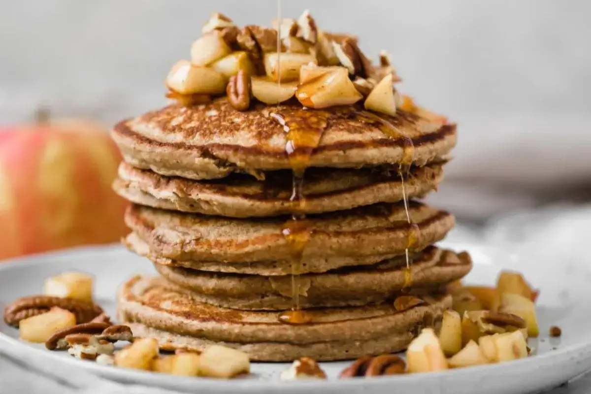 Stack of fluffy apple and oat pancakes with caramelized apples, maple syrup, and butter on a rustic plate.